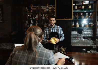 Smiling Bartender Talking to Customer at Counter. Young business woman in a formal wear sitting in a cafeteria at the bar after work and flirts with a bartender polishing glasses. - Powered by Shutterstock