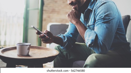Smiling bearded African man using smartphone while sitting on sofa at home.Concept people working with mobile gadget.Blurred background.Wide, crop