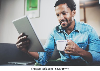 Smiling Bearded African Man Using Tablet For Reading Morning News And Drinking Black Coffee At Home.Concept People Working With Mobile Gadget.Blurred Background