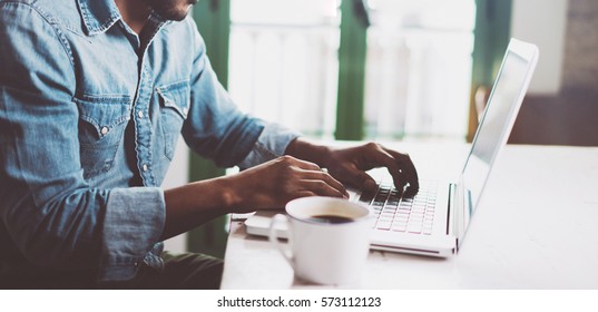 Smiling bearded African man using laptop at home while sitting the wooden table.Male hands typing on the notebook keyboard.Concept of young people work mobile devices.Blurred window background,wide.