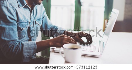 Photo of Smiling bearded African man using laptop at home while sitting the wooden table.Male hands typing on the notebook keyboard.Concept of young people work mobile devices.Blurred window background,wide.