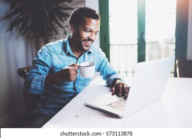 Smiling bearded African man working at home while sitting the wooden table.Using modern laptop for new job search.Concept of young people work mobile devices.Blurred background