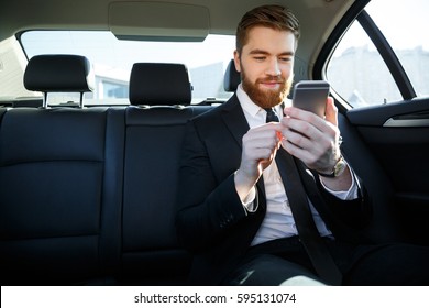 Smiling Bearded Business Man In Suit Looking At Mobile Phone In His Hand While Sitting In The Back Seat Of A Car