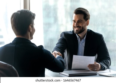 Smiling bearded businessman in suit shaking hands with job seeker. Happy successful male manager making deal with partner. Professional employee holding cv and greeting applicant.
