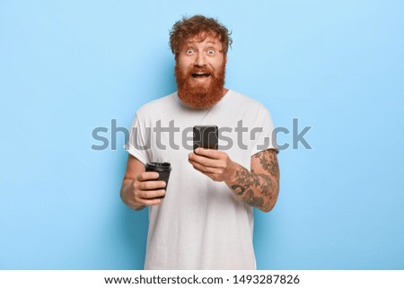 Smiling bearded emotional man has red hair, holds mobile phone, shares great news with friend, gazes at camera with broad smile and bugged eyes, wears casual white t shirt, holds takeaway coffee
