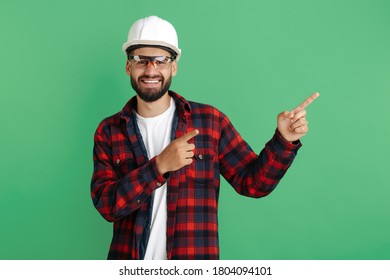 Smiling Bearded Engineer Or Constructor Man In Casual Outfit Pointing Finger Away Over Green Background.