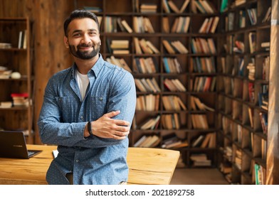 Smiling Bearded Indian Businessman Stands Near Desk And Looks At The Camera. Young Positive Male Student In Library With Bookshelves On Background. Proud And Successful Mixed-race Small Business Owner