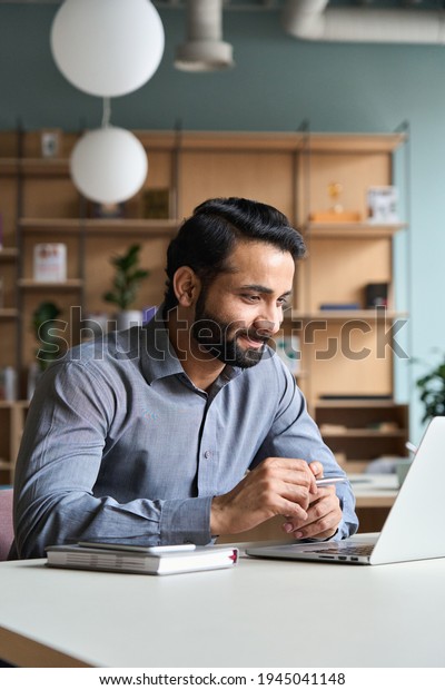 Smiling bearded indian businessman working on\
laptop at home office. Young indian student using computer remote\
studying, virtual training on video call meeting, watching online\
webinar or seminar.