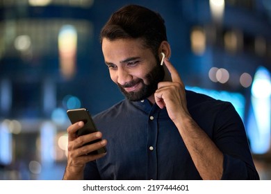 Smiling bearded indian man wearing earbud holding phone having video call at night. Eastern businessman in earphone using smartphone listening music in app tech on cellphone watching videos online. - Shutterstock ID 2197444601