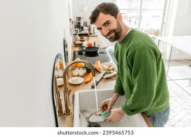Smiling bearded man in casual clothes looking at camera while washing plate at home - Shutterstock ID 2395465753