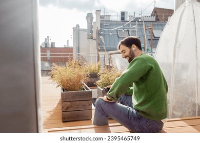 smiling bearded man in earphone and casual clothes using smartphone on house terrace rooftop - Shutterstock ID 2395465749