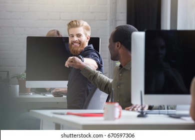Smiling bearded man giving a fist bump to a male colleague while they are sitting at their computer desks