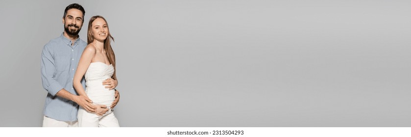 Smiling and bearded man hugging fashionable pregnant woman and looking at camera while standing isolated on grey with copy space, expecting parents concept, banner, baby bump, husband and wife - Shutterstock ID 2313504293