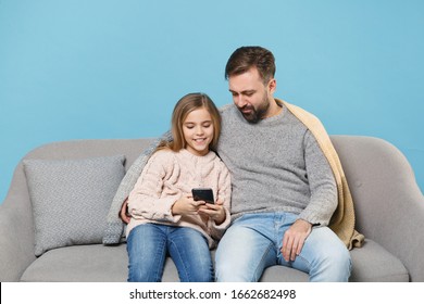 Smiling Bearded Man In Knitted Sweater With Child Baby Girl. Father Little Daughter Isolated On Pastel Blue Background. Love Family Parenthood Childhood Concept. Sitting On Couch, Hold Mobile Phone