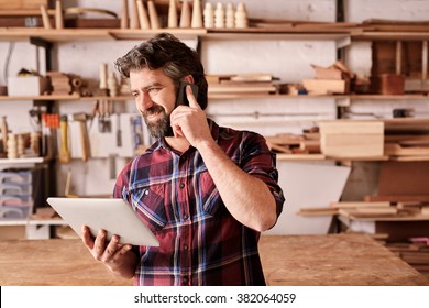 Smiling Bearded Man, Owner Of A Small Woodwork Business, Standing In His Workshop Studio, Talking On His Mobile Phone And Holding A Digital Tablet