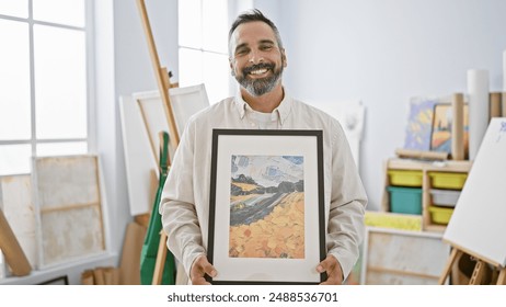 A smiling bearded man presenting a framed artwork in a bright artist's studio with easels and paintings. - Powered by Shutterstock