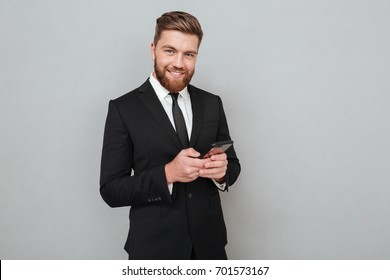 Smiling bearded man in suit using his smartphone and looking at the camera over gray background - Powered by Shutterstock