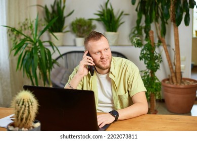 Smiling Bearded Man Talking Speaking On Phone Using Laptop. Male Employee Have Smartphone Call Cellphone With Client Or Business Partner.