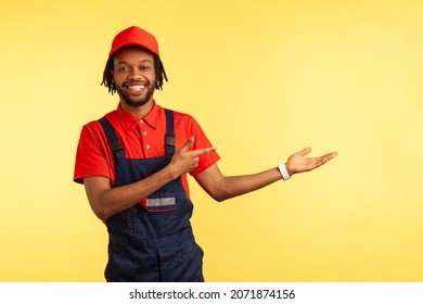 Smiling bearded mechanic wearing overalls presenting copy space on palm, pointing aside, advertisement of delivery or housekeeping service. Indoor studio shot isolated on yellow background.