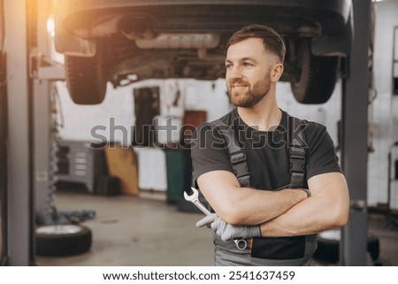 Smiling bearded mechanic while standing with crossed arms and holding wrenches in a car service