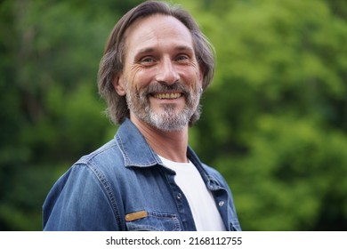 Smiling Bearded Middle Aged Man. Happy Old Man With Long Hair Walking In City Park. Portrait Of Man In Denim On Green Background