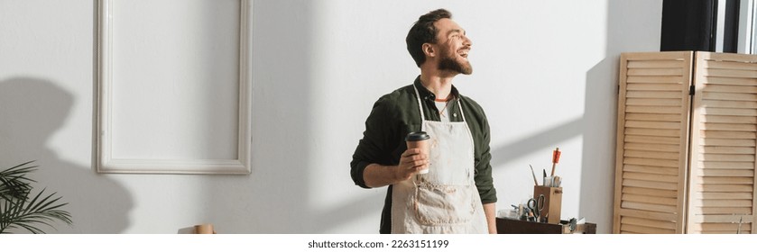Smiling bearded repairman in apron holding paper cup in workshop, banner - Shutterstock ID 2263151199