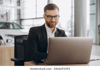 Smiling bearded salesman in suit and glasses typing on his laptop at new cars showroom - Powered by Shutterstock