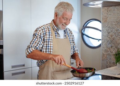 Smiling bearded senior man cooking vegetables holding a frying pan. People in the kitchen preparing dinner. An elderly man alone cooking at home - Powered by Shutterstock