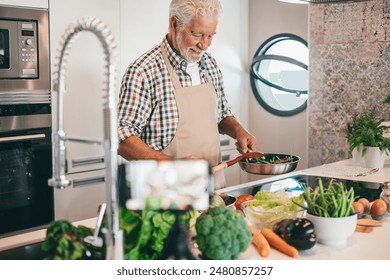 Smiling bearded senior man holding frying pan with vegetables. People in the kitchen preparing dinner. An elderly man alone cooking at home - Powered by Shutterstock