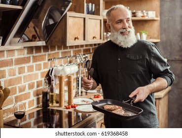 Smiling bearded senior man holding frying pan with steaks and looking away in kitchen - Powered by Shutterstock