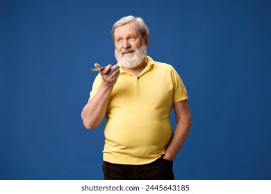 Smiling, bearded senior man in yellow polo recording voice message via mobile phone against blue studio background. Easy communication. Concept of human emotions, lifestyle, casual fashion - Powered by Shutterstock