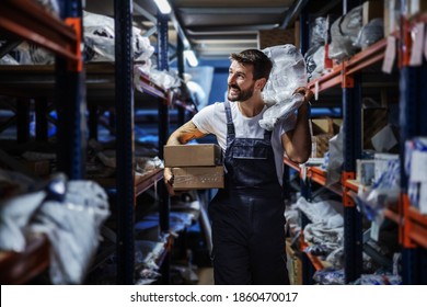 Smiling Bearded Tattooed Hardworking Blue Collar Worker In Overalls Holding Boxes And Bag And Relocating Them While Walking In Storage Of Import And Export Firm.