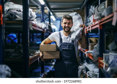 Smiling Bearded Tattooed Hardworking Blue Collar Worker In Overalls Holding Boxes And Bag And Relocating Them While Walking In Storage Of Import And Export Firm.