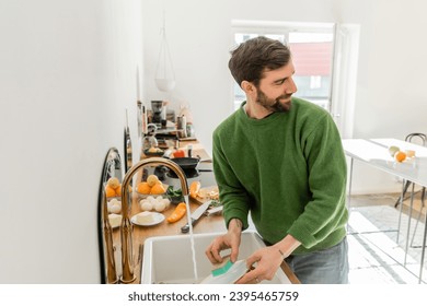 Smiling bearded and tattooed man washing plate near sink and looking away at home - Shutterstock ID 2395465759