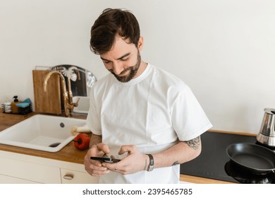 Smiling and bearded tattooed man in white t-shirt using smartphone while standing near worktop - Shutterstock ID 2395465785