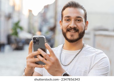 Smiling bearded young man using smartphone typing text messages browsing internet, finishing work, looking at camera outdoors. Guy tourist walking in urban sunshine city street. Town lifestyles
