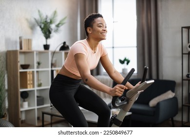 Smiling beautiful african american sports woman in sportswear cycling bike at home on background of light living room. Cardio training, exercising legs, cardio workout indoors. - Powered by Shutterstock