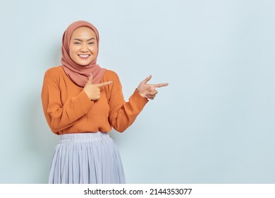 Smiling beautiful asian woman in brown sweater and hijab pointing finger in copy space, showing advertising products and confident looking at camera isolated over white background.
