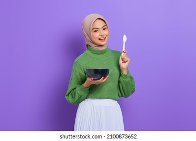 Smiling beautiful Asian woman in green sweater and hijab holding spoon and bowl with cereal isolated over purple background - Powered by Shutterstock