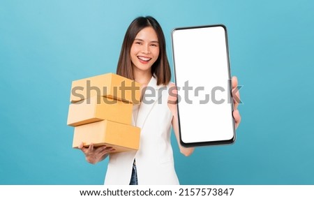 Smiling beautiful Asian woman holding cardboard boxes and hands show smartphone mockup of blank screen on blue background. Take your screen to put on advertising. Concept delivery online.