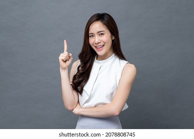 Smiling beautiful Asian woman pointing finger upward to empty space in studio gray isolated background