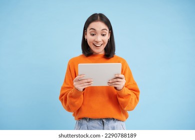 Smiling beautiful asian woman reading on digital tablet, watching videos on gadget or playing video games, standing over blue background.