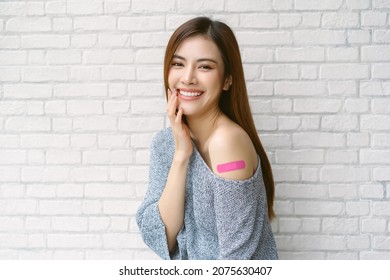 Smiling beautiful Asian woman wearing off shoulder sweater sleeve showing her arm with adhesive bandage plaster after receiving the Covid-19 Vaccine on white brick wall background. Vaccine Safe lives.