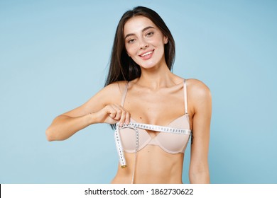 Smiling beautiful attractive young brunette woman 20s in beige brassiere underwear standing posing measuring breast with measure tape isolated on pastel blue colour wall background studio portrait