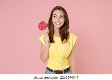 Smiling beautiful attractive young brunette woman 20s wearing yellow casual t-shirt posing standing holding in hands round lollipop looking aside isolated on pastel pink background studio portrait