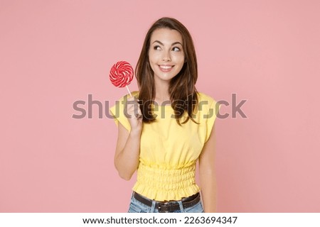Smiling beautiful attractive young brunette woman 20s wearing yellow casual t-shirt posing standing holding in hands round lollipop looking aside isolated on pastel pink background studio portrait