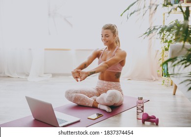 Smiling beautiful attractive young strong sporty fitness woman wearing pink tracksuit doing yoga exercises using laptop computer sitting and stretching hands on mat floor at home indoor