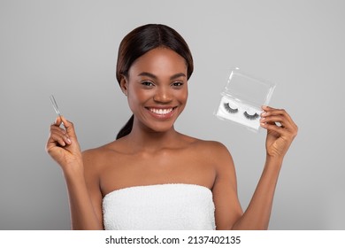 Smiling beautiful black woman wrapped in towel holding pack of false eyelashes and tweezers while posing on grey background, happy young african american female ready to apply artificial eye lashes