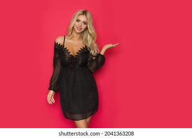 Smiling Beautiful Blonde Woman In Black Lace Dress Is Holding Hand Raised And Presenting. Front View. Three Quarter Length Studio Shot On Red Background.