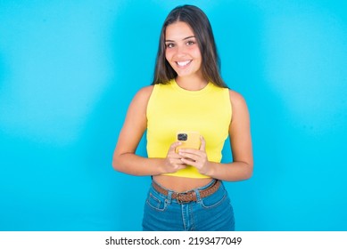 Smiling Beautiful Brunette Woman Wearing Yellow Tank Top Over Blue Background Friendly And Happily Holding Mobile Phone Taking Selfie In Mirror.